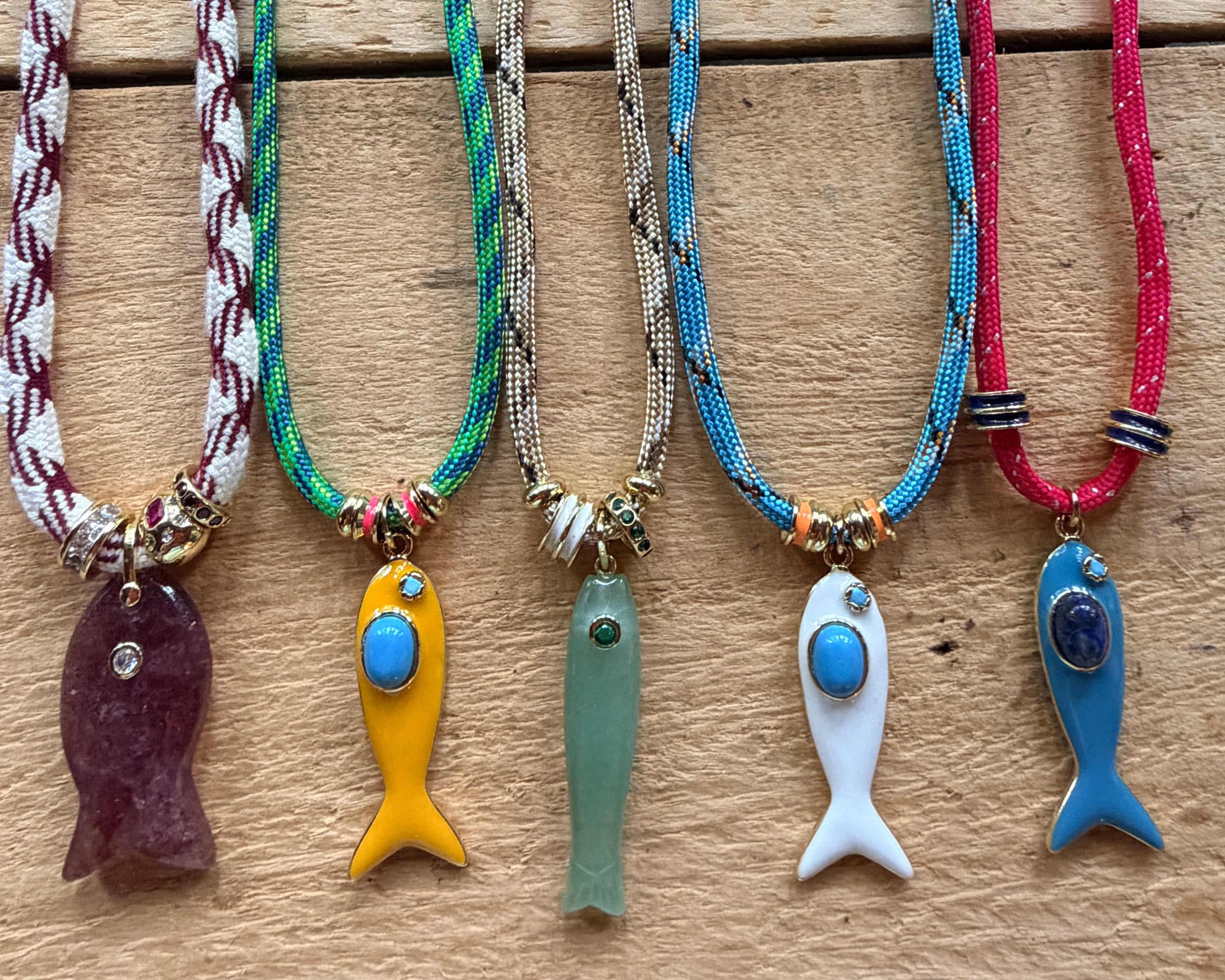 Five handmade beaded fish necklaces with colorful cords and fish pendants are displayed on a light wood background.